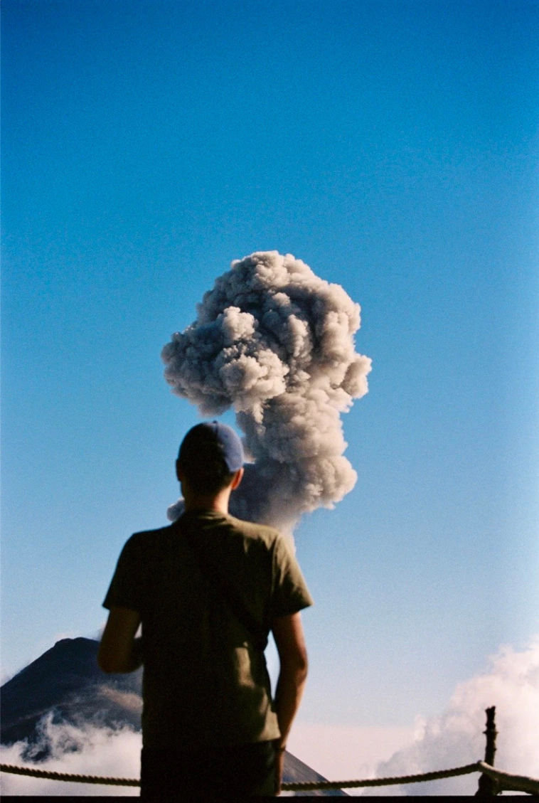 Silhouette d’un homme de dos, les fumées volcaniques s’échappant et prolongeant sa forme, sous un ciel bleu.